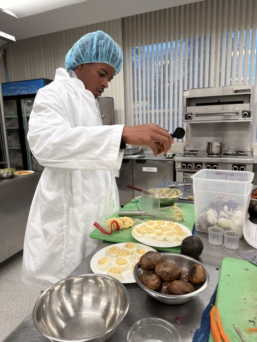 A person in a food preparation area wearing a white coat and blue hair net is adding an ingredient to a dish on a counter filled with ingredients. There are sliced fruits and vegetables, and a piece of avocado on nearby plates. Kitchen equipment is visible in the background.