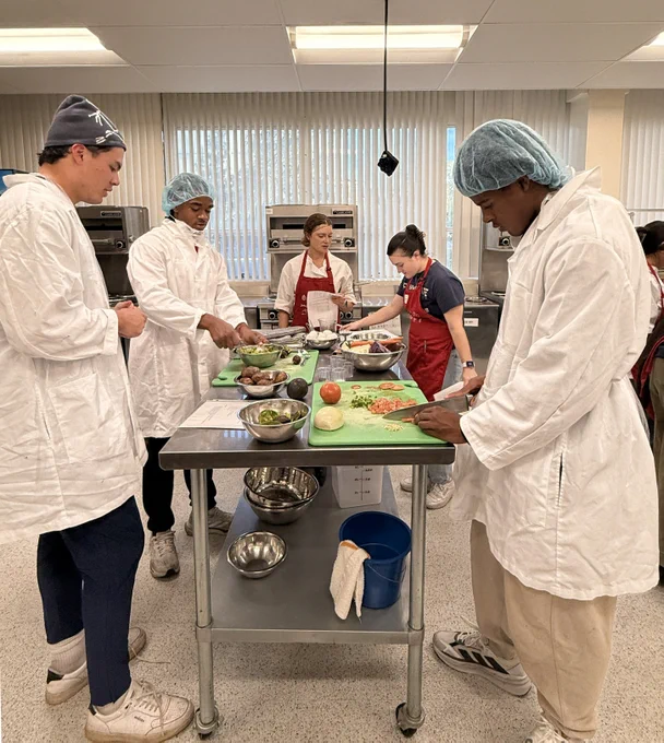A group of people in a kitchen setting is preparing food. They are wearing white lab coats and protective hair coverings. Various vegetables and ingredients are on the countertops, along with mixing bowls and utensils. The environment appears to be a professional kitchen or cooking class.