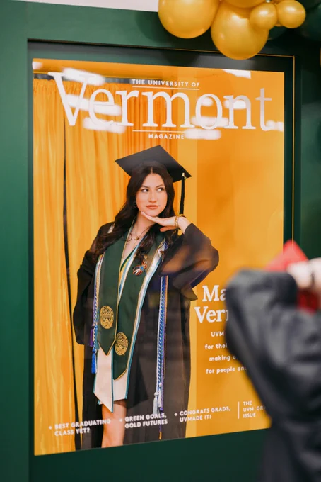 This is an image of a large poster featuring a person in a graduation cap and gown, posed thoughtfully with one hand under their chin. They are wearing a green stole with emblems. The poster is titled "Vermont" and is part of "The University of Vermont Magazine." Balloons are visible at the top of the poster.