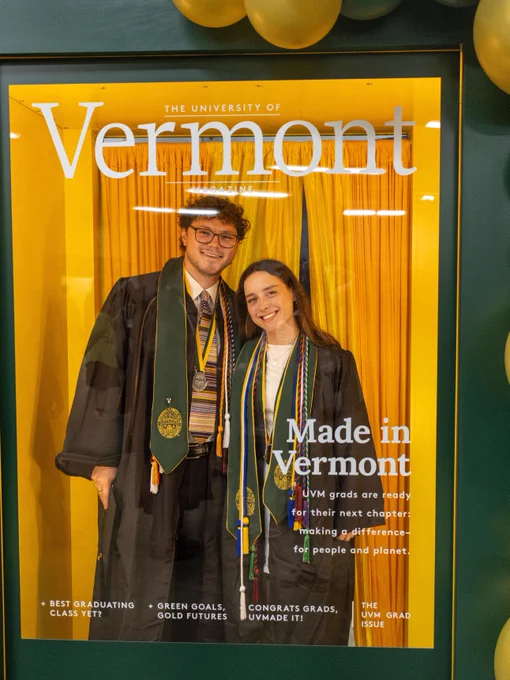Two individuals in graduation attire are smiling and posing in front of a backdrop for The University of Vermont. The backdrop includes congratulatory messages and balloons.