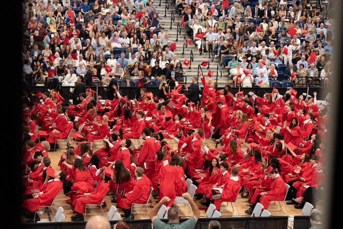 A large crowd of people wearing red graduation gowns and caps seated in rows, with a backdrop of a packed audience in a theater or auditorium setting.