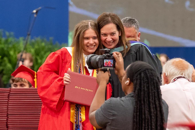Two smiling young women in graduation gowns and caps, one holding a diploma, posing for a photo with a camera operator.