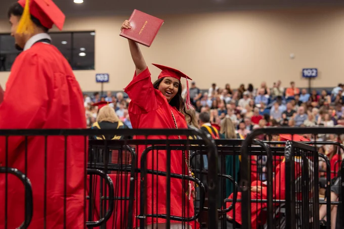 A graduate in a red graduation gown stands behind a metal railing, holding up their diploma and smiling amid a crowd of other graduates.