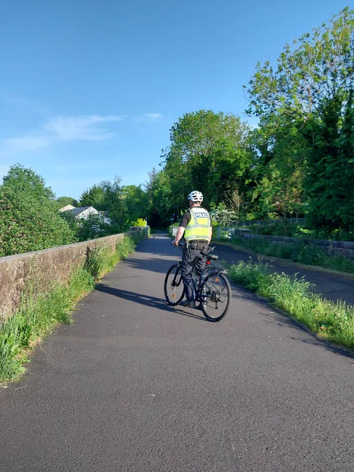 A police officer on a bicycle wearing a high-visibility vest and helmet, cycling on a paved path.