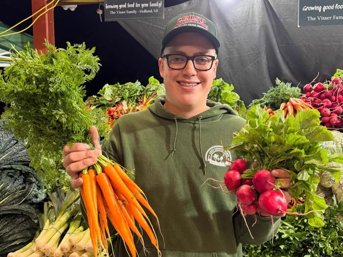 A person smiles while holding a bunch of fresh carrots in one hand and radishes in the other at a market stall. They are wearing a cap and a green hoodie. The background shows more vegetables and signs reading "Growing good food for your family.