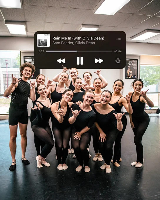 A group of ballet dancers pose together in a dance studio, all smiling and making a friendly hand gesture. A music overlay shows the track "Rein Me In (with Olivia Dean)" by Sam Fender and Olivia Dean.