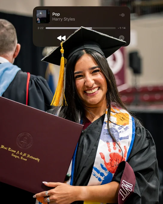 A person in graduation attire is smiling and holding a diploma cover. They are wearing a decorated graduation stole. In the background, blurry figures in similar attire can be seen. At the top, there's a music player overlay showing "Pop" by Harry Styles.