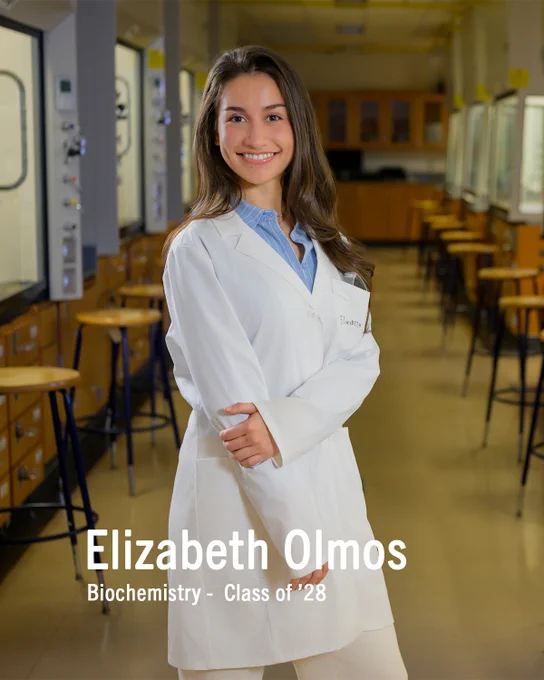 A person in a lab coat stands confidently in a science laboratory with rows of benches and equipment. The text reads: "Elizabeth Olmos, Biochemistry - Class of '28."