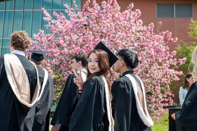 Graduates wearing black gowns and caps celebrate outdoors near a flowering tree. One person turns back toward the camera with a peace sign. A modern building is visible in the background.