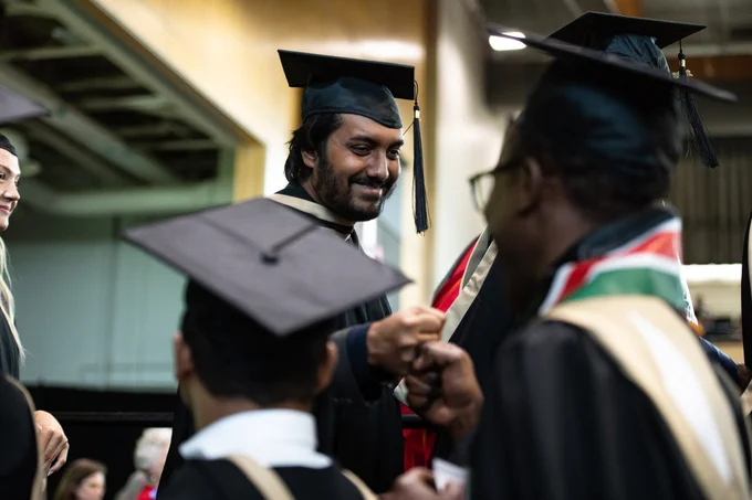A group of people wearing graduation caps and gowns are celebrating. Two individuals are fist-bumping, while others are standing nearby, smiling and chatting. The setting is an indoor graduation ceremony.