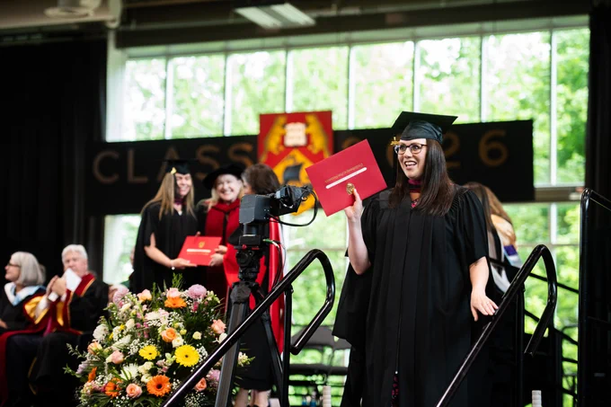 A person in graduation attire holds a red diploma cover while smiling towards the camera. They are on a stage decorated with a floral arrangement and a backdrop that says "Class of 2026." Seated individuals in academic robes are visible in the background.