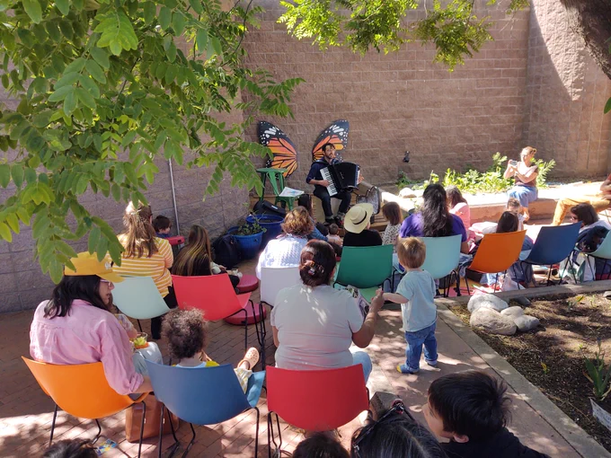Musician Jordan Wax plays accordion in the garden in front of an audience of parents and children sitting in colorful chairs.