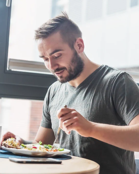 A person seated at a table enjoys a fresh salad from a plate with a fork. The setting is bright with natural light coming through a large window.