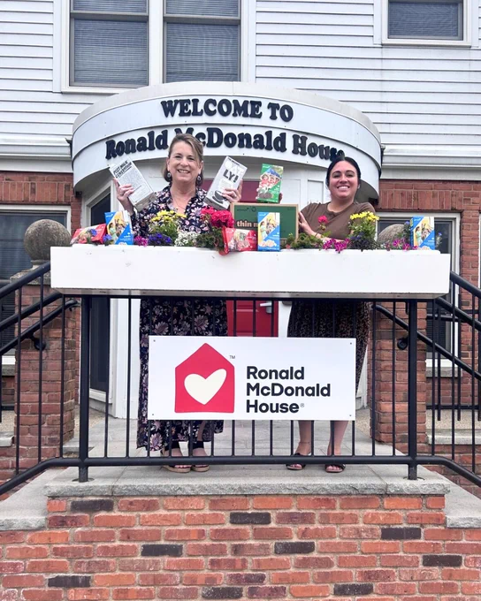 Two people stand on a porch in front of Ronald McDonald House. They hold various items and are surrounded by small plants and flowers. A "Ronald McDonald House" sign with a heart logo is visible on the railing.
