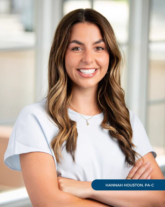 Hannah Houston, PA-C, of Tryon Medical Partners smiles with her arms crossed in a professional portrait posed in front of a paned glass window