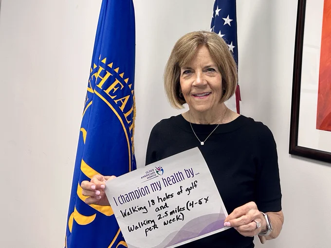 Mary Lazare, ACL’s Principal Deputy Administrator, stands in an office holding a sign that reads, “I champion my health by… walking 18 holes of golf and walking 2.5 miles (4-5x per week).” An HHS flag and a U.S. flag are visible in the background.
