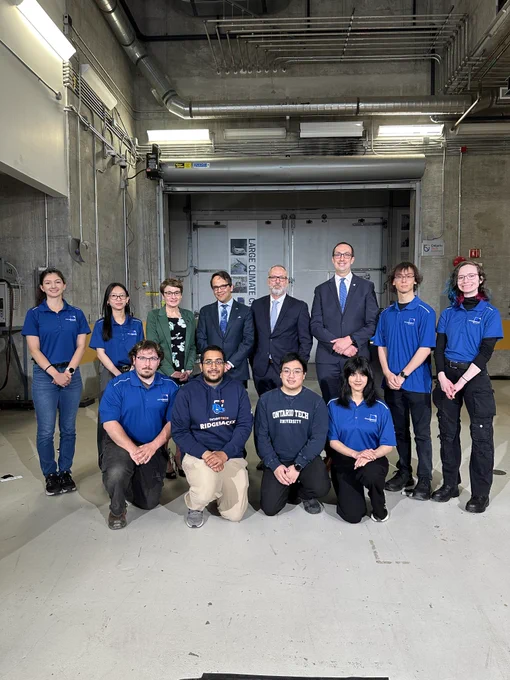 Dr. Steven Murphy, President and Vice-Chancellor of Ontario Tech University, Dr. Elaine Popp, President of Durham College, Daniel Tisch, President and CEO of the Ontario Chamber of Commerce, and Hon. Nolan Quinn, Minister of Colleges, Universities, Research Excellence and Security (MCURES) pose for a group photo with a group of Ontario Tech students dressed in blue Ontario Tech branded shirts inside the university's ACE facility.
