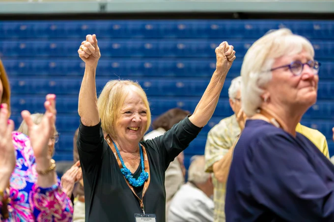 A woman raises both arms in celebration while surrounded by a crowd in an indoor venue with blue bleachers.