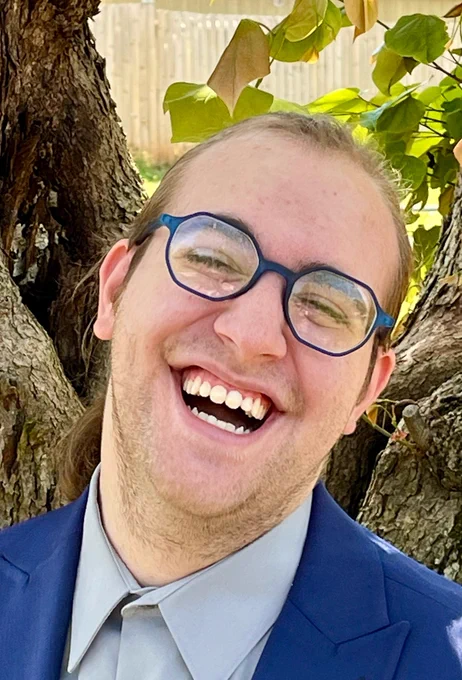 Derek stands in front of a tree wearing a navy suit, grey shirt, and blue hexagonal glasses. Their eyes are squinted and they have an open-mouthed smile, as if they’ve been caught mid-laugh.