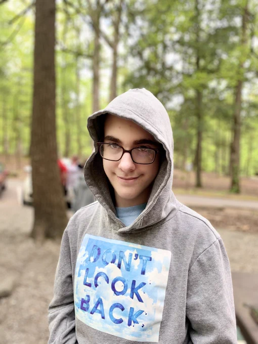 Derek stands in a wooded area with square glasses and a hoodie that reads “Don’t Look Back.” They have a closed mouth half-smirk and a bit of facial hair.