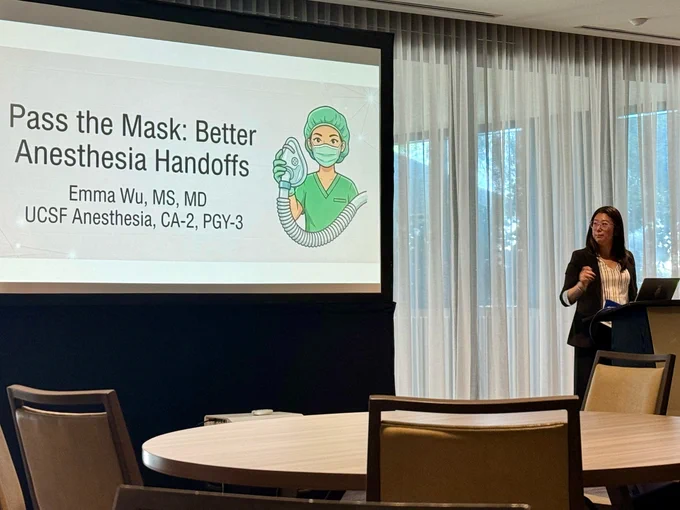 A woman wearing a suit and glasses standing at a lectern and looking at a large projector screen that reads, "Pass the Mask: Better Anesthesia Handoffs, Emma WU, MS, MD, UCSF Anesthesia, CA-2, PGY-3."