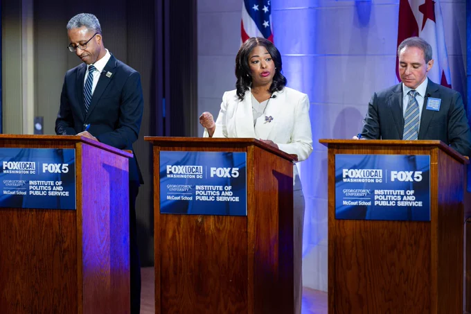 a man in black suit, a woman in white suit, and a man in gray suit stand at three lecterns