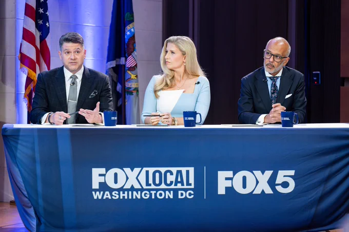 a man in black suit, a woman in light blue suit, and a man in navy suit sit at a Fox 5 table