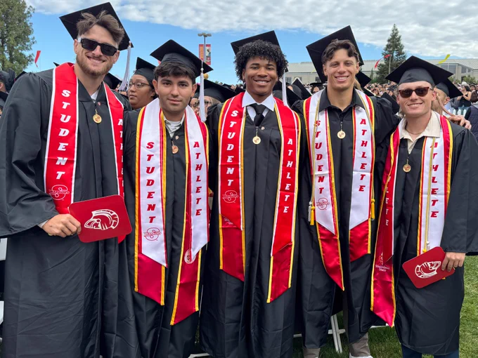 Five Stan State graduates stand together at commencement wearing black caps and gowns with red-and-white “Student Athlete” stoles while smiling for a group photo.