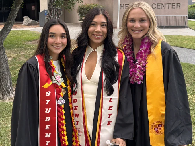Three Stan State graduates pose together outdoors in graduation attire, wearing stoles, leis, and cords during commencement celebrations.