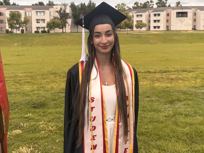 A Stan State graduate wearing a black cap and gown, honor cords, and a “Student Athlete” stole poses outdoors on campus during commencement.