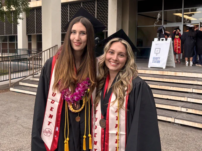 Two Stan State graduates wearing caps and gowns smile together outside a campus building while wearing medals, leis, cords, and “Student Athlete” stoles.