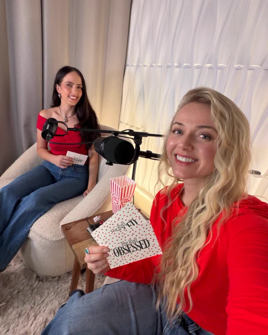 Two women in red tops record a podcast in a cozy studio set up with microphones and cards. The mood is friendly and engaging.