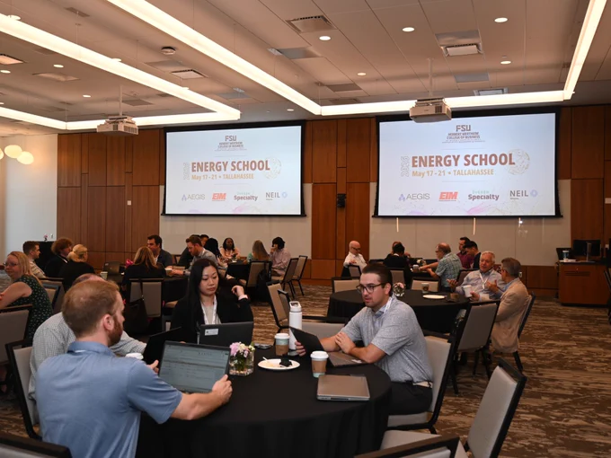 A conference room with several round tables where people are engaged in discussions. Two large screens display "FSU Energy School, May 17-21, Tallahassee" with logos for AEGIS, IRM, Specialty, and NEIL.