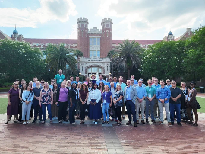 A group of people are posing for a photo outside a large red-brick building with two towers at Florida State University. The scene includes palm trees and a partly cloudy sky in the background.
