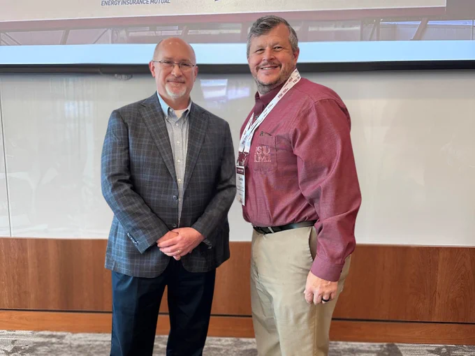 Two people are standing side by side in a professional setting. One is wearing a plaid suit jacket and glasses, while the other is wearing a maroon shirt with the Texas State University logo. Both are smiling and standing in front of a presentation screen in a conference room.