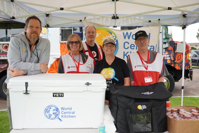A group of five people stands under a canopy featuring a "World Central Kitchen" banner. They are gathered around a table with a YETI cooler displaying the World Central Kitchen logo. Two individuals wear Red Cross vests. There are food supplies visible, including jars and a black bag. A food truck is partially visible in the background.