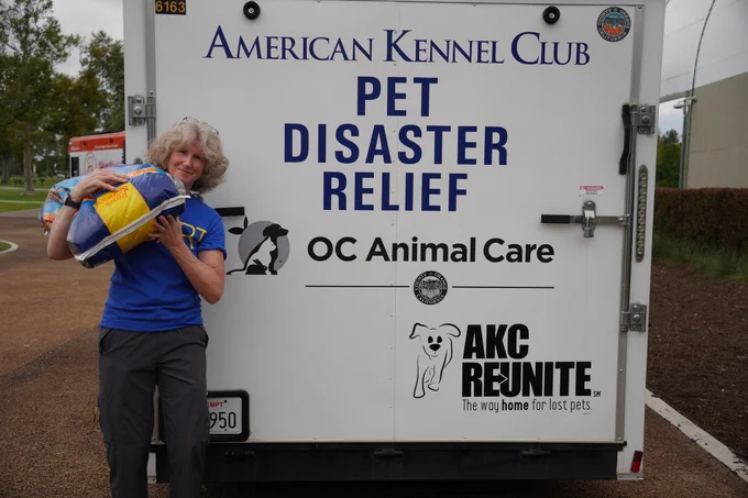 A person stands smiling next to a trailer labeled "American Kennel Club Pet Disaster Relief" with logos for "OC Animal Care" and "AKC Reunite." The person holds cat litter. The trailer is parked outdoors.