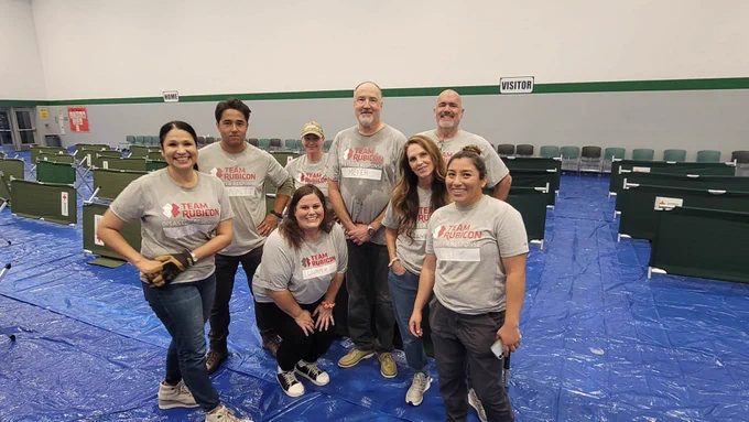 A group of people wearing matching "Team Rubicon" t-shirts pose together in a room with green cots set on a blue tarp floor. 