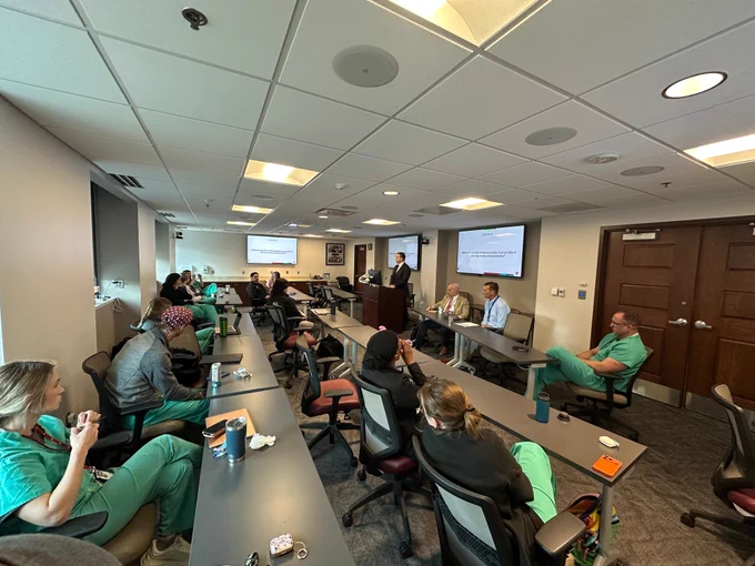 A group of medical professionals and trainees sit in a conference room during a Grand Rounds presentation. A speaker stands at the podium while panelists sit at the front of the room. Presentation slides about healthcare policy are displayed on screens as attendees in scrubs listen and participate in the discussion.