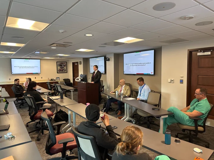 A group of medical professionals and trainees sit in a conference room during a Grand Rounds presentation. A speaker stands at the podium while panelists sit at the front of the room. Presentation slides about healthcare policy are displayed on screens as attendees in scrubs listen and participate in the discussion.