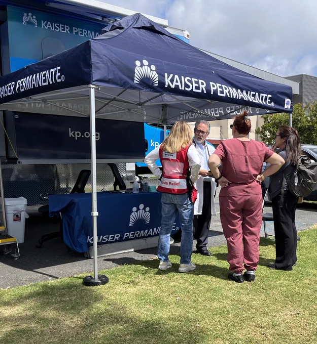 A group of people standing under a Kaiser Permanente tent next to a mobile unit. Two wear uniforms, and one has a badge. The scene takes place outdoors on grass.