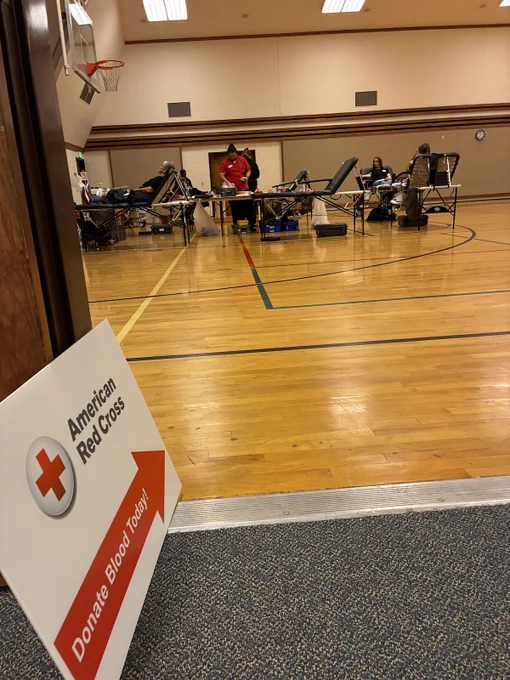 Gym with blood donation spots set up, and a sign with the Red Cross logo on it. Below the logo is a red arrow pointing into the gym with "Donate blood here" written inside the arrow.