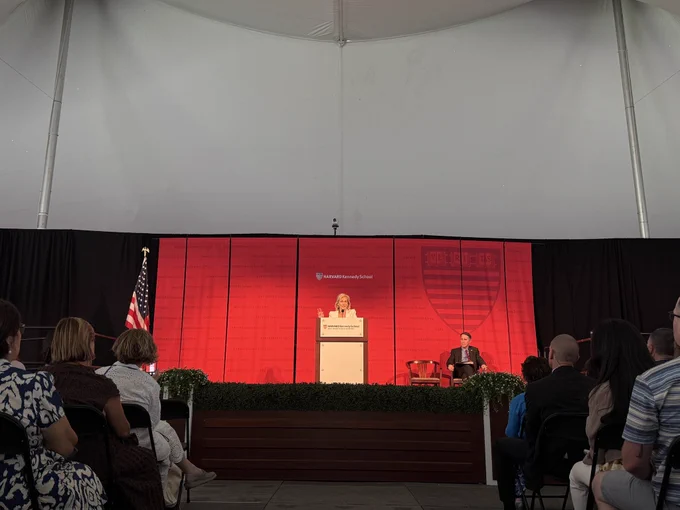 Zanny Minton Beddoes giving the 2026 address to graduates on a stage and podium with a red Harvard Kennedy School backdrop behind her.