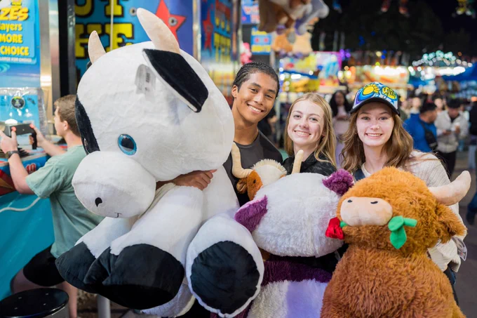 Two young women and one young man standing on the midway at the N.C. State Fair holding three stuffed animal prizes. 