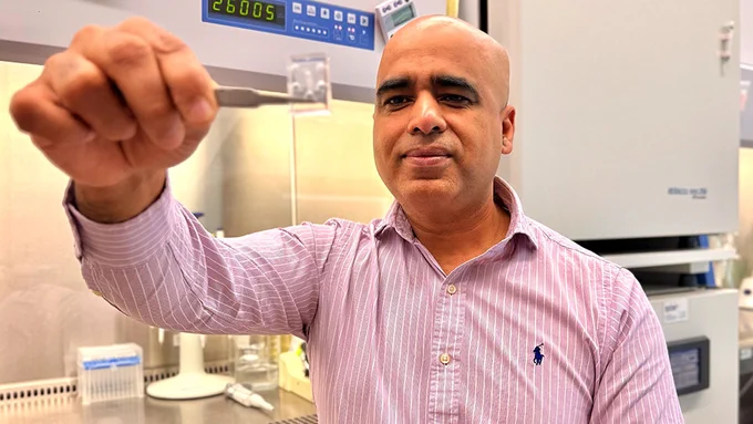 A male researcher uses tweezers to hold a plastic chip toward the camera. He sits in a lab in front of a fume hood.