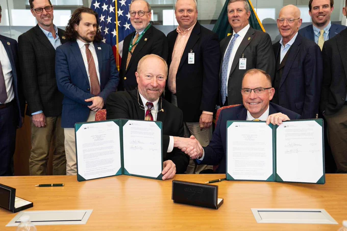 Chief Shultz of the forest service shakes hands and holds up an MOU with various men and women in the background. 