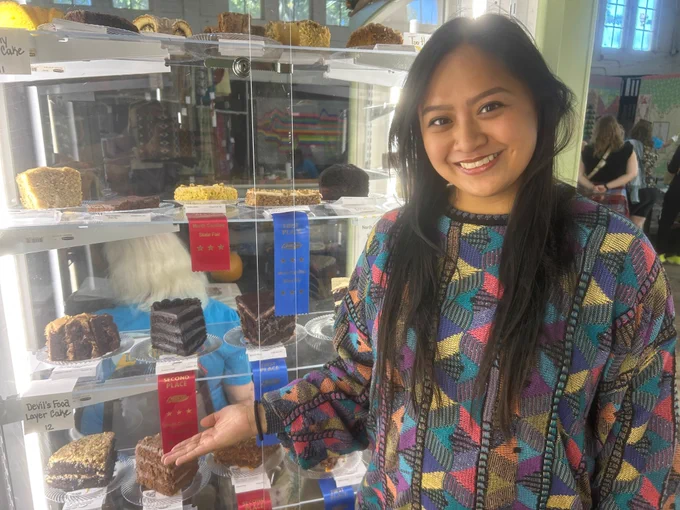 A young Asian girl standing in front of a glass case full of baked goods and pointing to a piece of cake with a red second-place ribbon on it. 