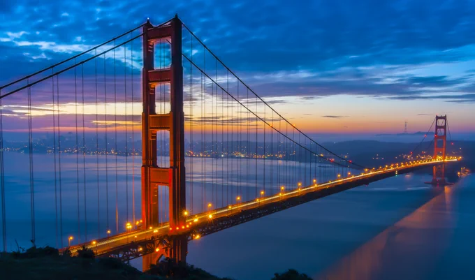 Golden Gate Bridge illuminated at dusk with city lights and a colorful sky in the background.