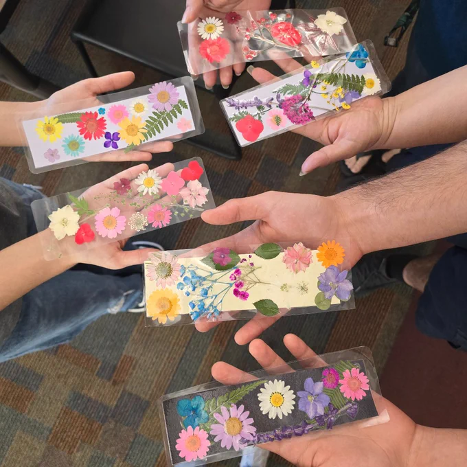Six hands holding rectangular bookmarks embedded with colorful pressed flowers over a patterned carpet floor.