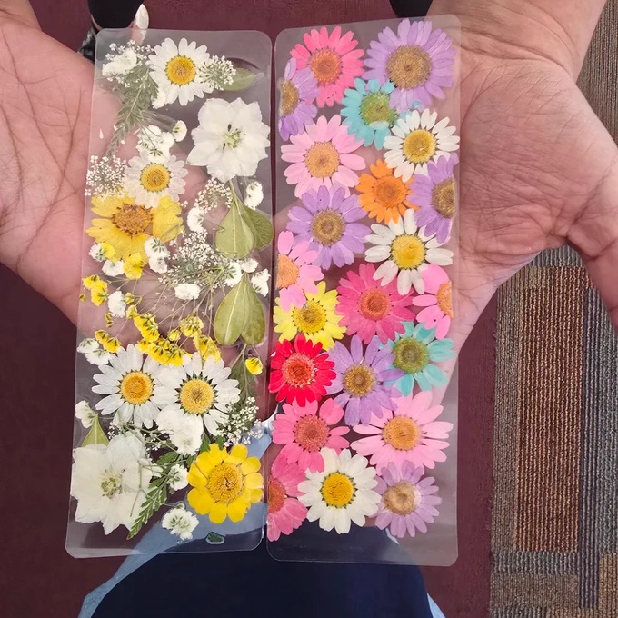 Two clear bookmarks filled with pressed flowers in white, yellow, pink, orange, and purple hues held by two hands.